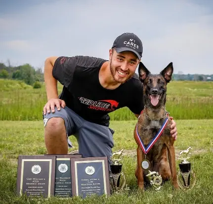 Blair Lowe kneeling beside his Belgian Malinois, Zira, with a medal, trophies and award plaques from a PSA competition.